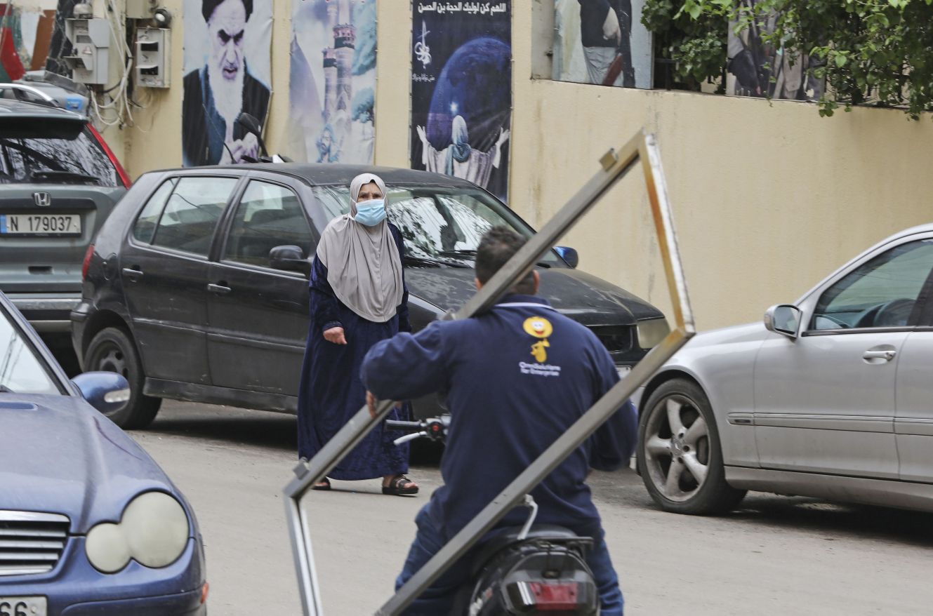 A woman, wearing a face mask amid coronavirus fears, crosses a street with a portrait of Iran’s former Supreme Leader Ayatollah Ruhollah Khomeini hanging on the wall in Beirut’s southern suburb of Chiyah on 14 March 2020. (Credit: Getty Images)